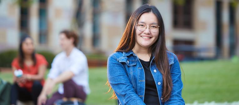 Student sitting on a bench with a grassy area and sandstone building in the background.